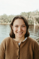 Lindsey Headshot Photo, She is wearing a brown cardigan and smiling into the camera. There is a warm backdrop of a lake behind her.