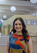 Cristina Headshot, she is smiling into the camera and wearing a multicolored top. She is indoors with light bulbs in the background and frames on the wall.